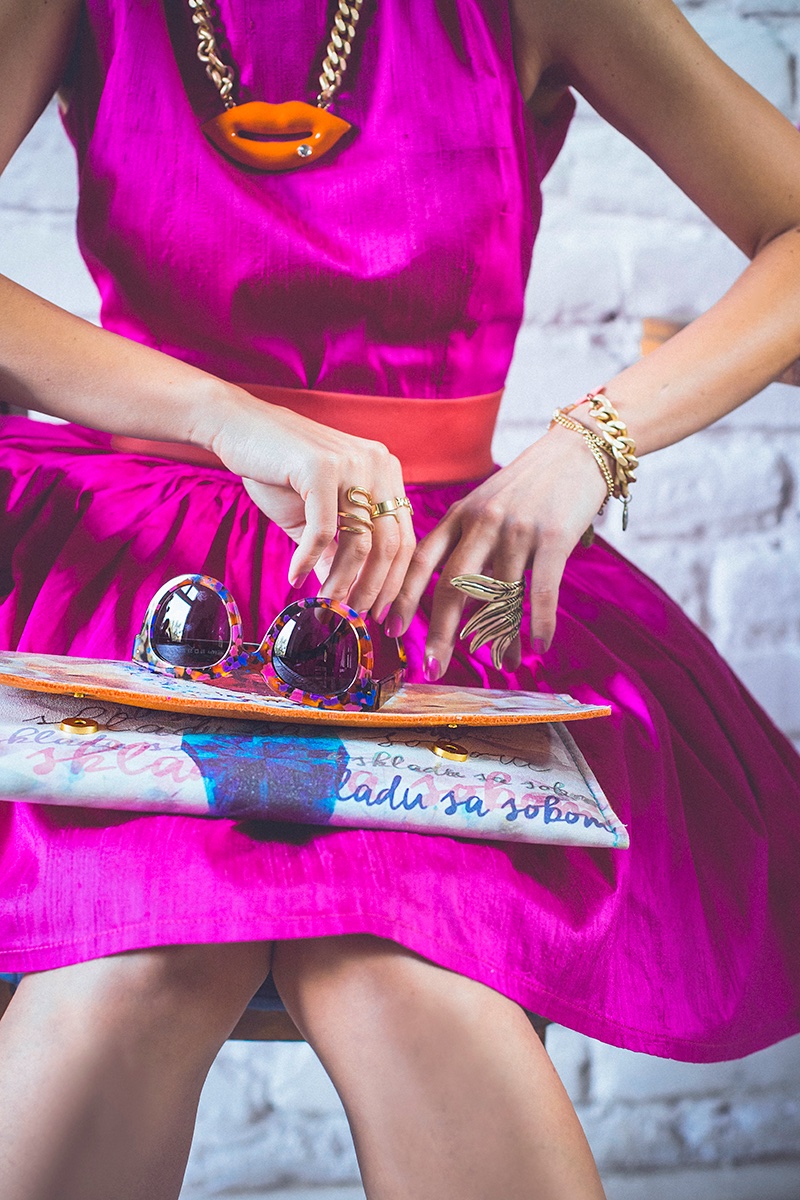 Detalj stila — naočare za sunce, ogrlica u obliku usana i šarena torbica u krilu