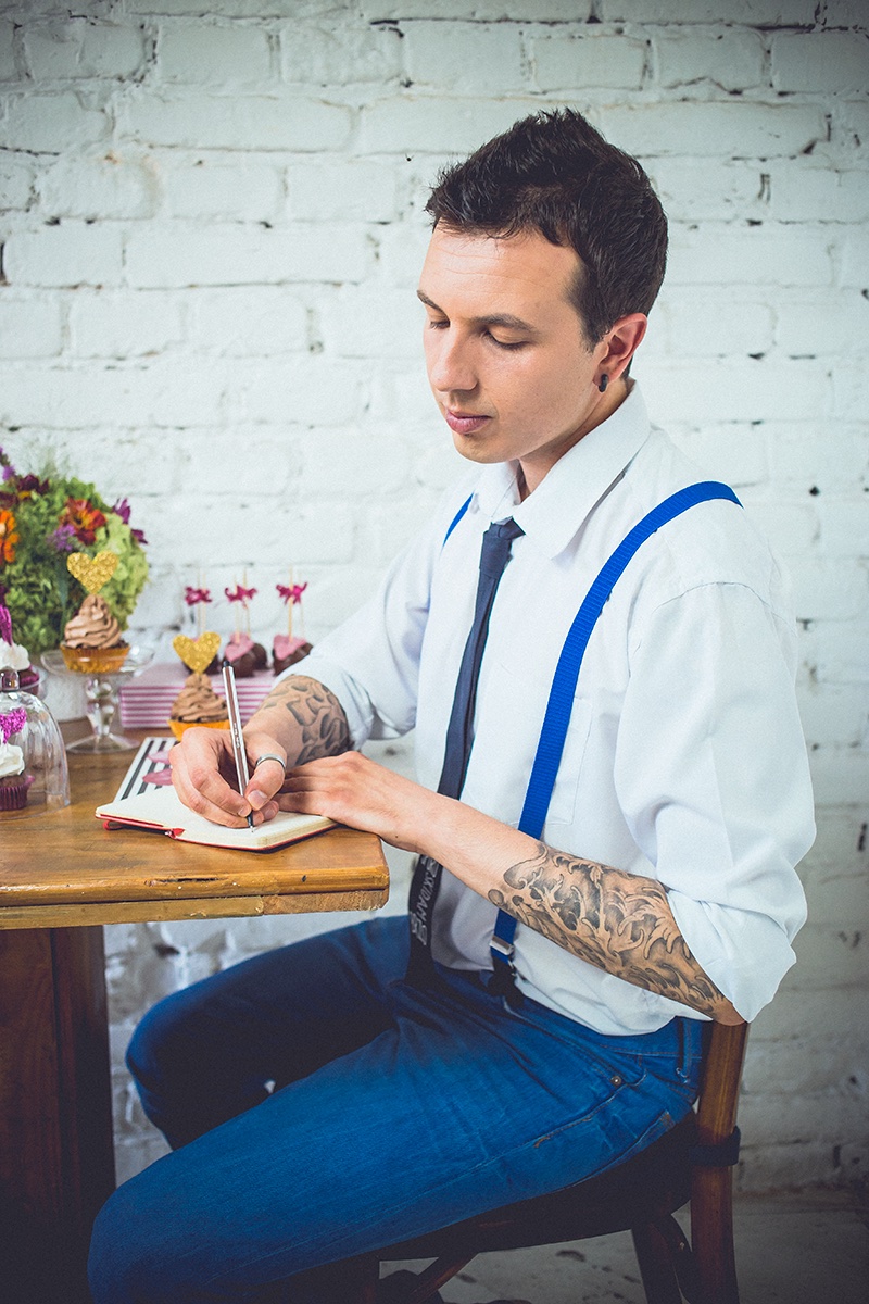 Marko piše u notes za stolom — tetovaže, tregeri i kapkejkovi u pozadini