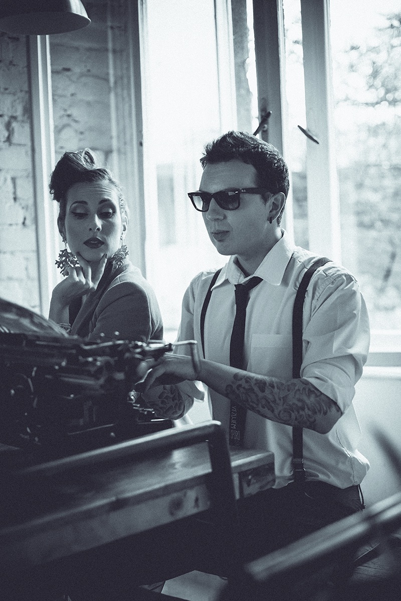 Crno-beli portret para uz vintage pisaću mašinu — on sa tetovažama i tregerima