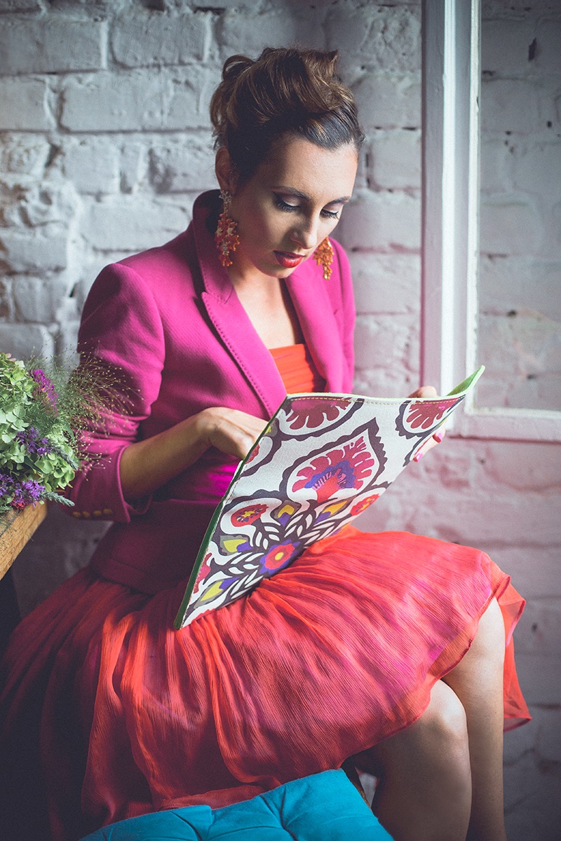 Ona sedi kraj prozora sa folk-print torbicom — magenta sako i koralna suknja