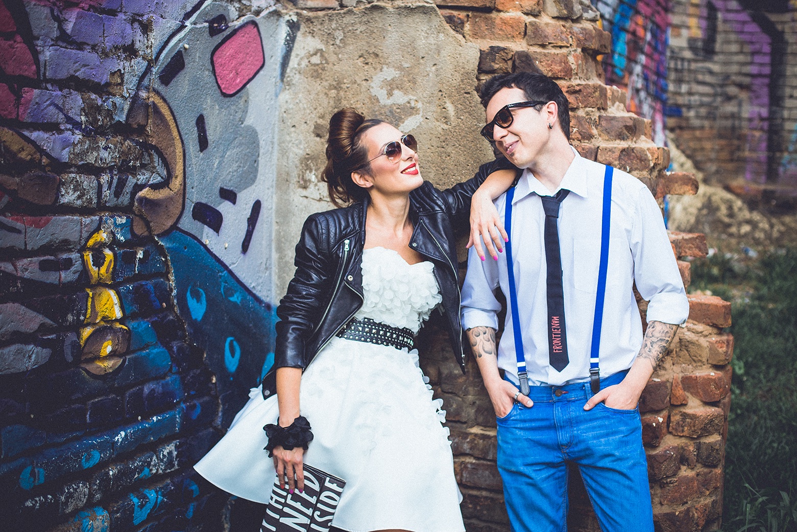 Par se smeje uz grafitni zid — kožna jakna, bela haljina, tregeri i naočare