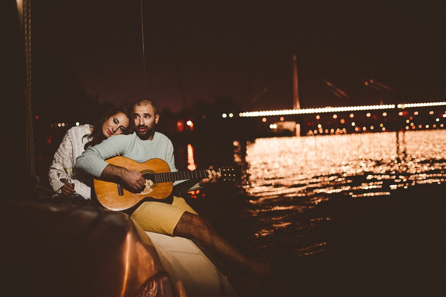 Gitara na jedrilici — noćni kadar, svetla grada na vodi