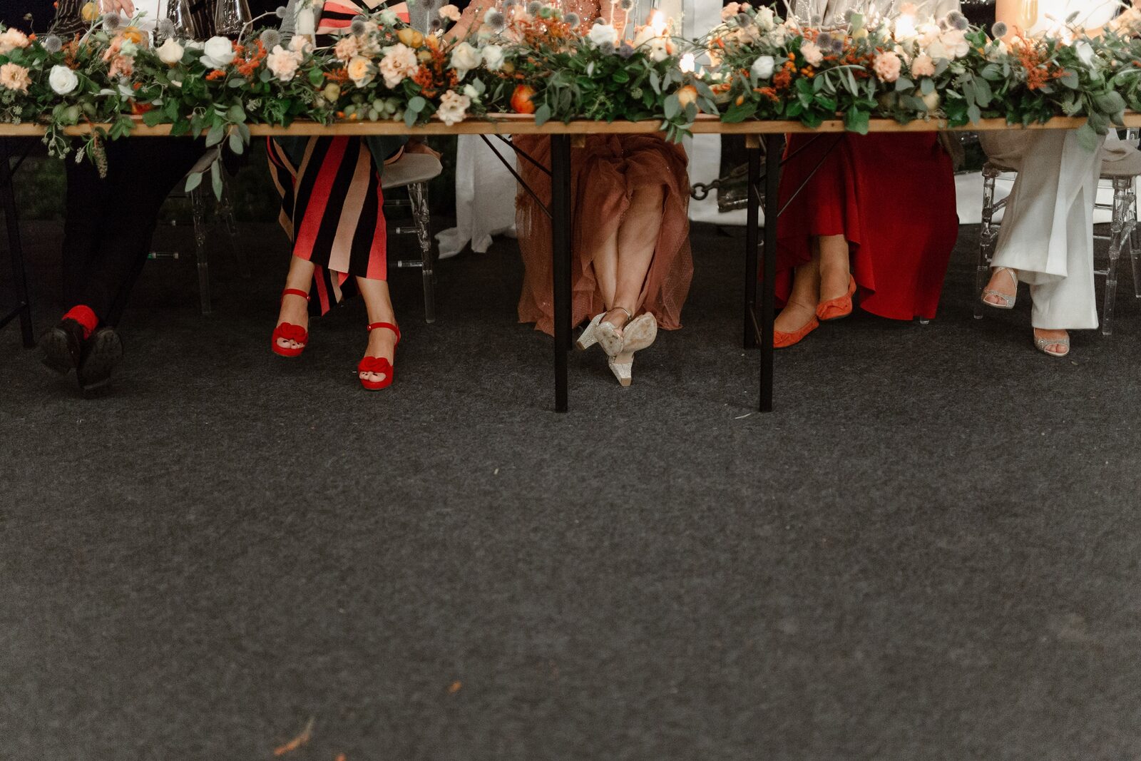 Noge gostiju ispod stola — crvene sandale, elegantne haljine